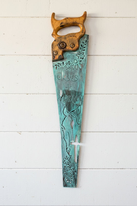 A decorative wall saw with a turquoise patina blade engraved with 'Jesus + Love' and floral motifs, and a weathered brown handle.
