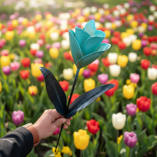 A hand holding a decorative metal tulip garden flower with turquoise-blue petals and black leaves against a background of tulips.
