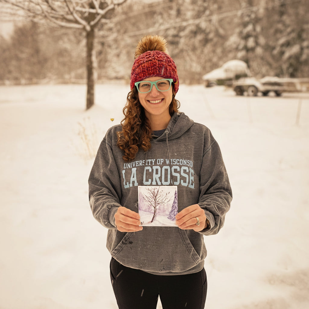 A woman, the artist, standing in a snowy outdoor setting, wearing a red knit beanie, glasses, a gray University of Wisconsin La Crosse hoodie, and black pants, while holding a small square painting of the maple tree behind her.