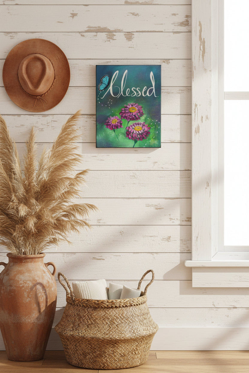 A rectangular canvas painting with the word 'Blessed' in white script over a blue and green painted background, with three pink-purple zinnia flowers and a blue butterfly.