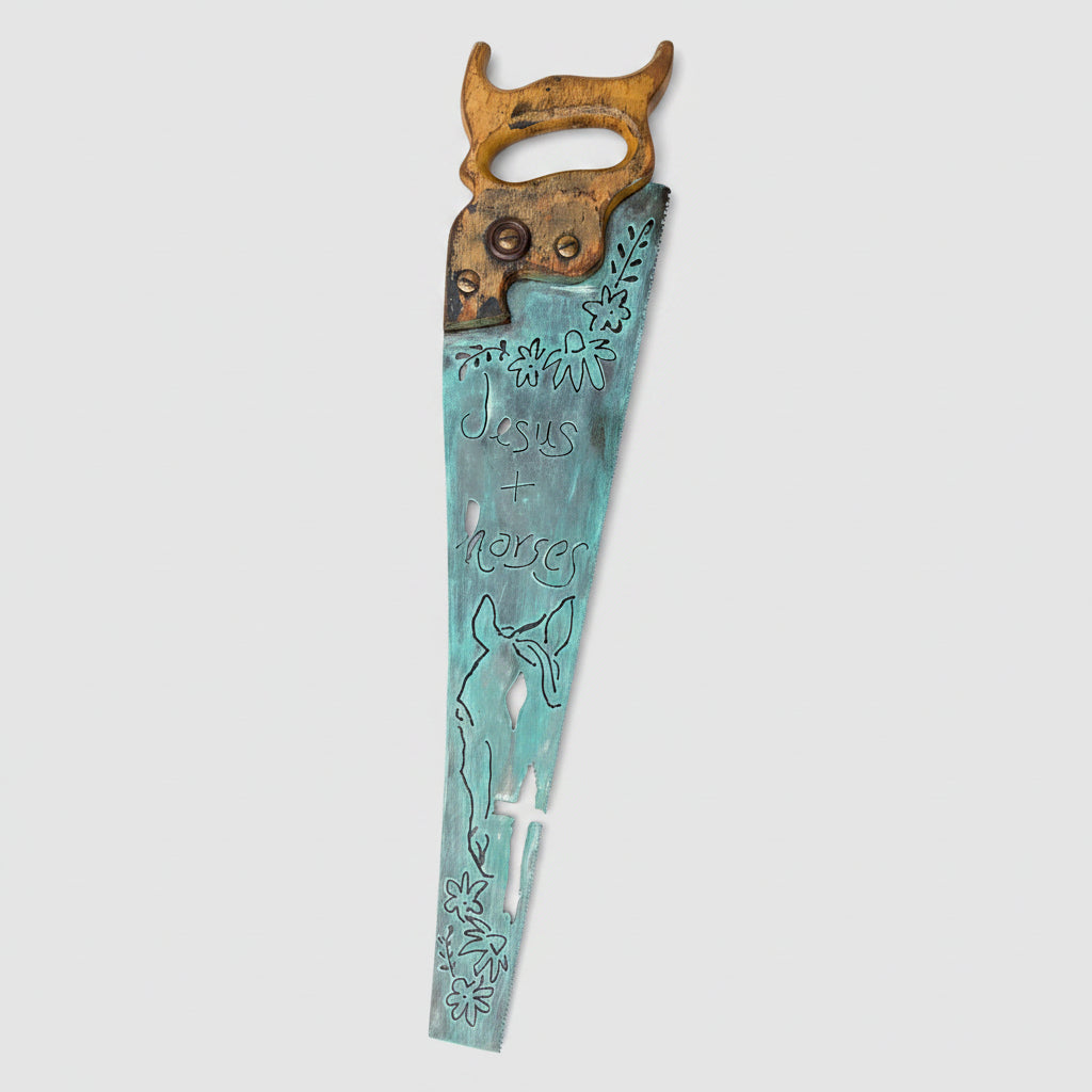 A rustic metal wall sign created from a vintage hand saw, stained in a distressed turquoise blue. The words 'Jesus + horse'. The wood handle is brown, and the sign is displayed against a gray wood-paneled background.