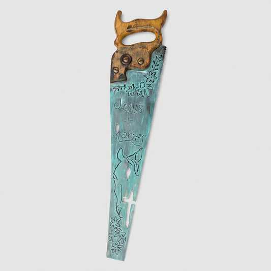 A rustic metal wall sign created from a vintage hand saw, stained in a distressed turquoise blue. The words 'Jesus + horse'. The wood handle is brown, and the sign is displayed against a gray wood-paneled background.