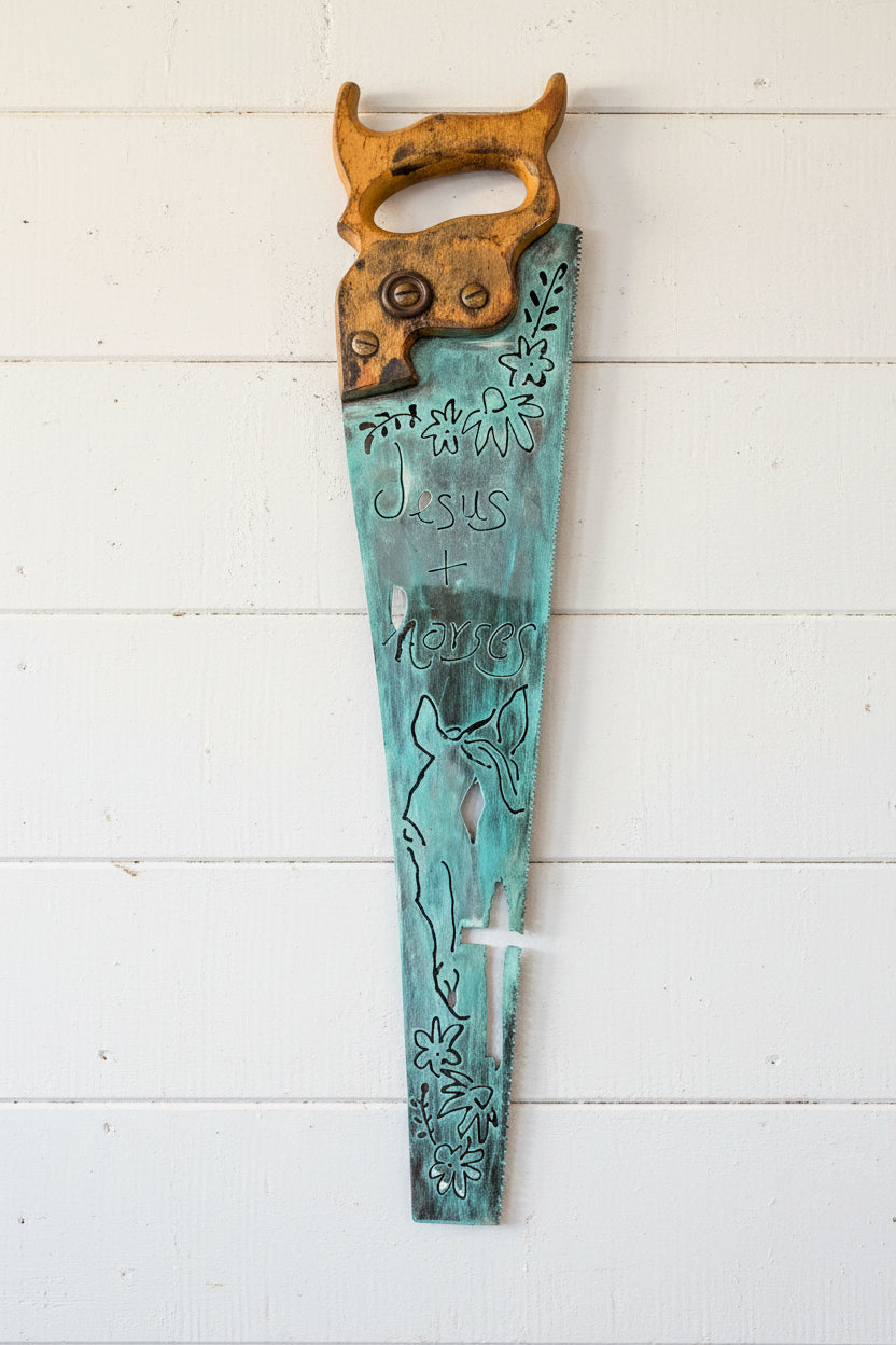 A decorative wall saw with a turquoise patina blade engraved with 'Jesus + Love' and floral motifs, and a weathered brown handle.