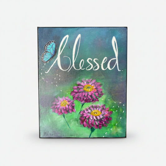 A rectangular canvas painting featuring the word 'Blessed' in white script over a blue and green painted background, with three pink-purple zinnia flowers and a blue butterfly.