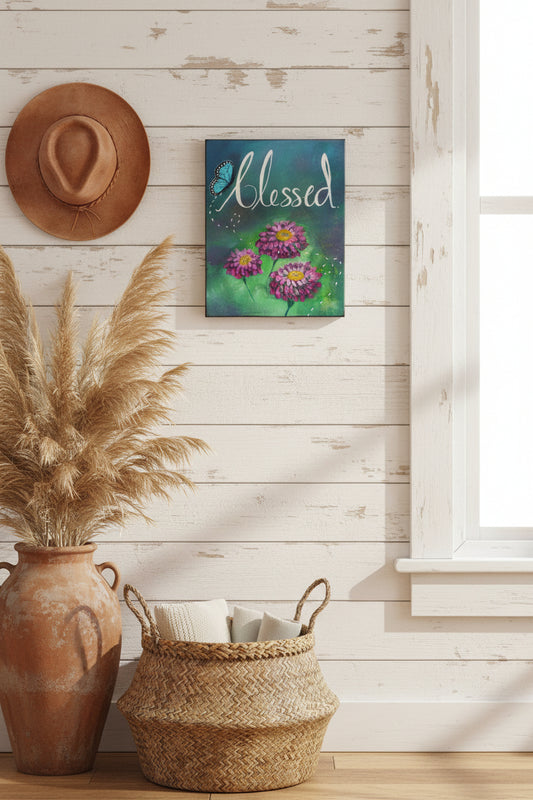 A rectangular canvas painting with the word 'Blessed' in white script over a blue and green painted background, with three pink-purple zinnia flowers and a blue butterfly.