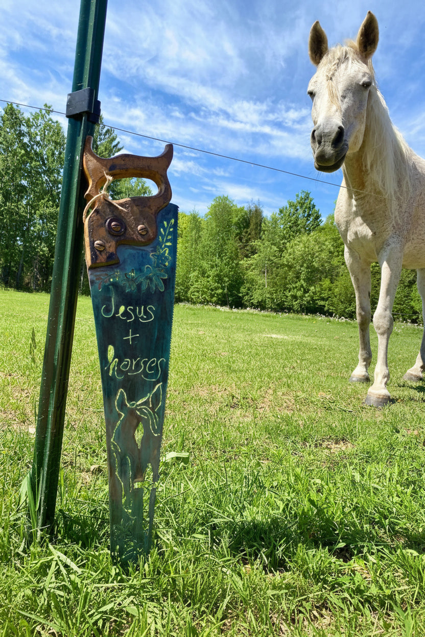 An old decorative handsaw leaning against a t-post in a grassy field, featuring carved words 'Jesus + horses.' A white horse stands in the background under a blue sky.