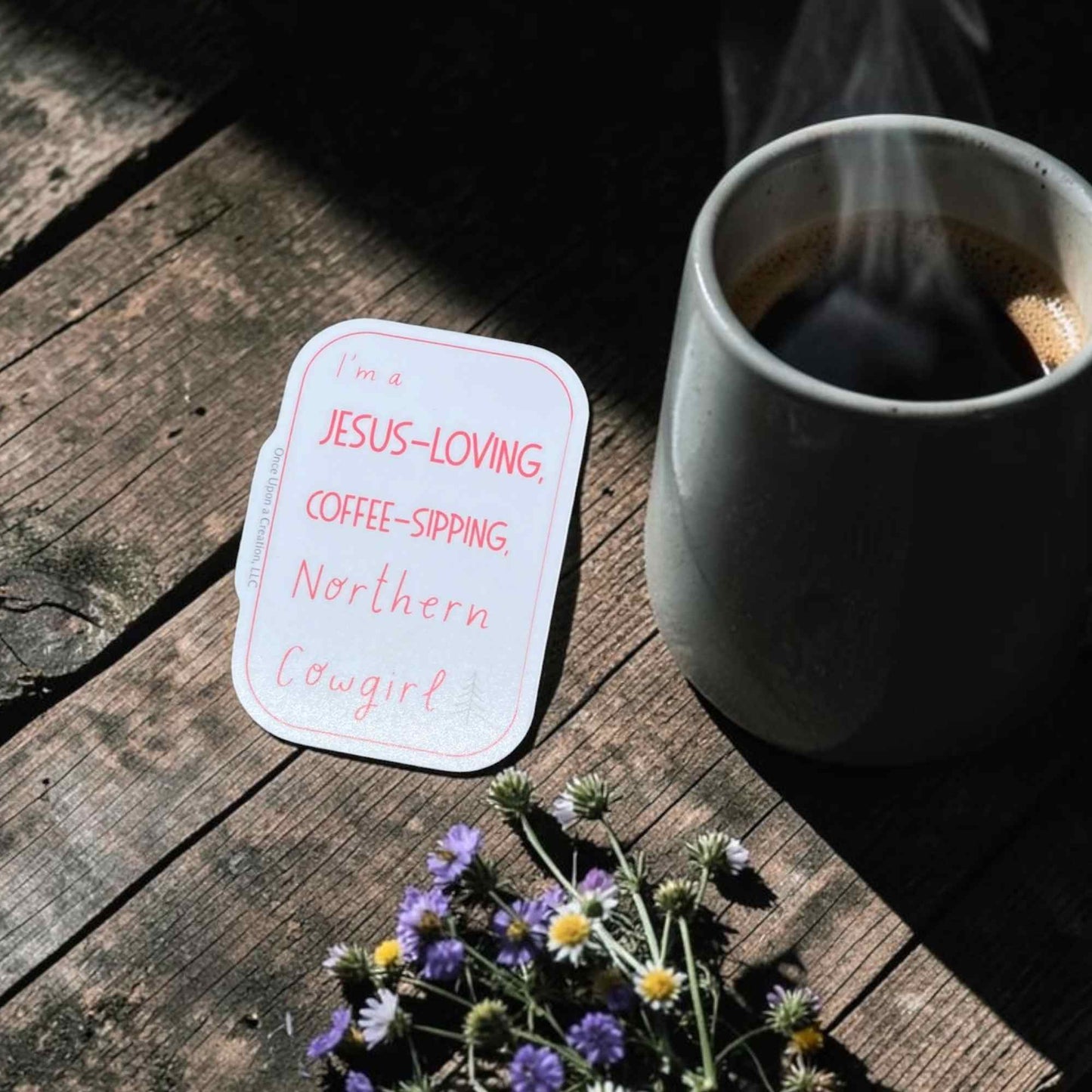 A die-cut white sticker with coral text reading 'I'm a JESUS-LOVING, COFFEE-SIPPING, Northern Cowgirl' placed on a rustic wooden table next to a gray ceramic mug of coffee and small purple wildflowers.