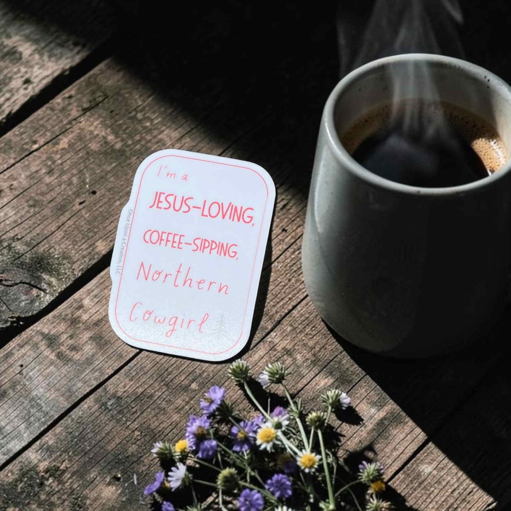 A die-cut white sticker with coral text reading 'I'm a JESUS-LOVING, COFFEE-SIPPING, Northern Cowgirl' placed on a rustic wooden table next to a gray ceramic mug of coffee and small purple wildflowers.