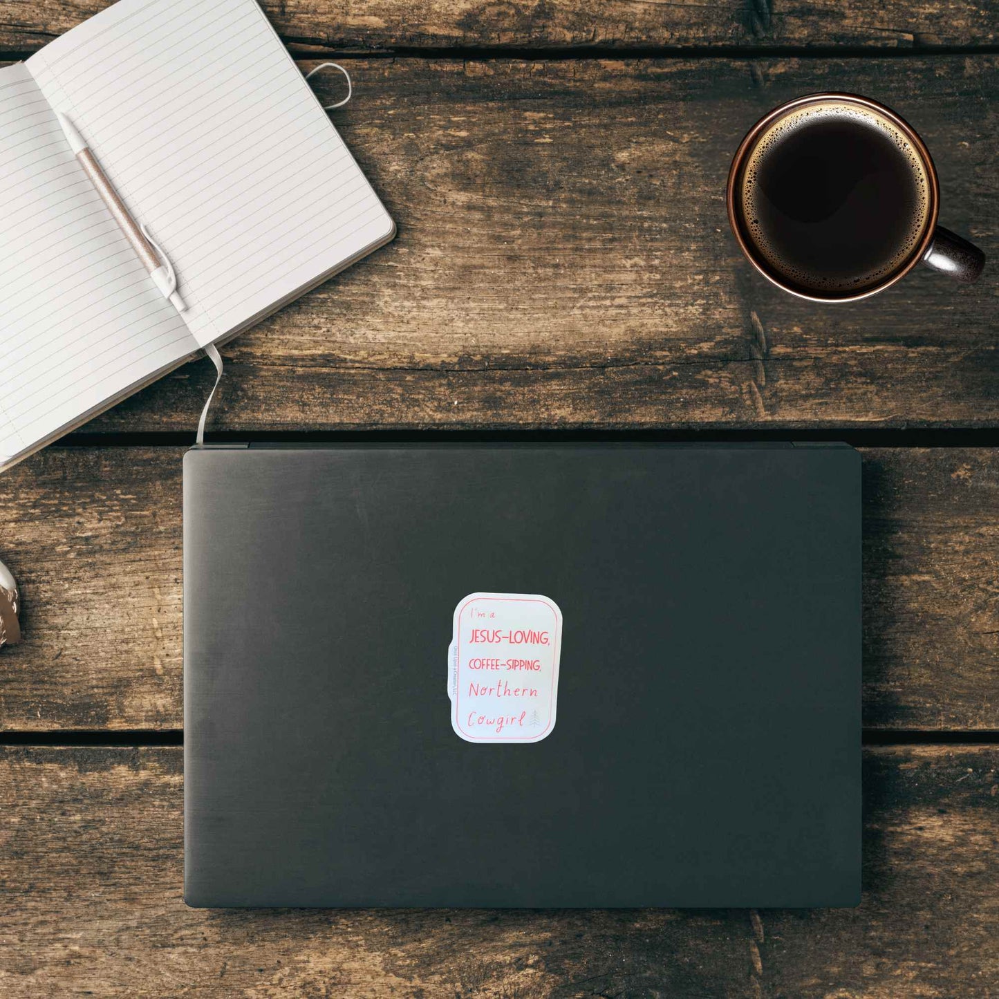 A black laptop with a white sticker that reads 'I'm a Jesus-loving coffee-sipping northern cowgirl' lies on a rustic wooden table, styled with an open notebook and a cup of coffee.
