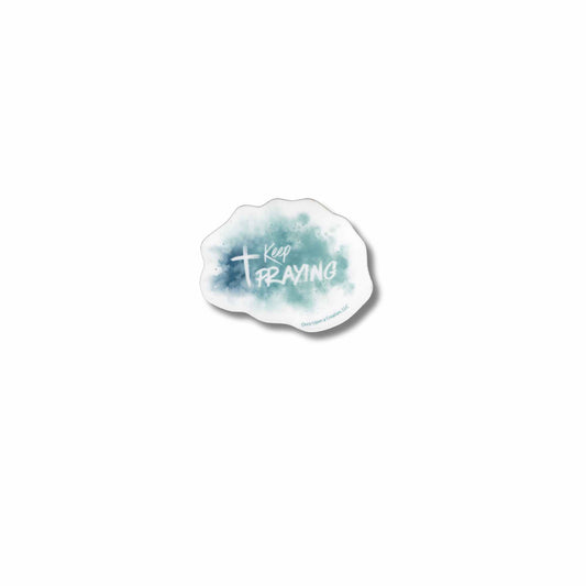 A die-cut sticker with a white background and a blue-green watercolor wash, featuring the text 'Keep PRAYING' with a white cross and a small 'Once Upon a Creation, LLC' logo at the bottom.