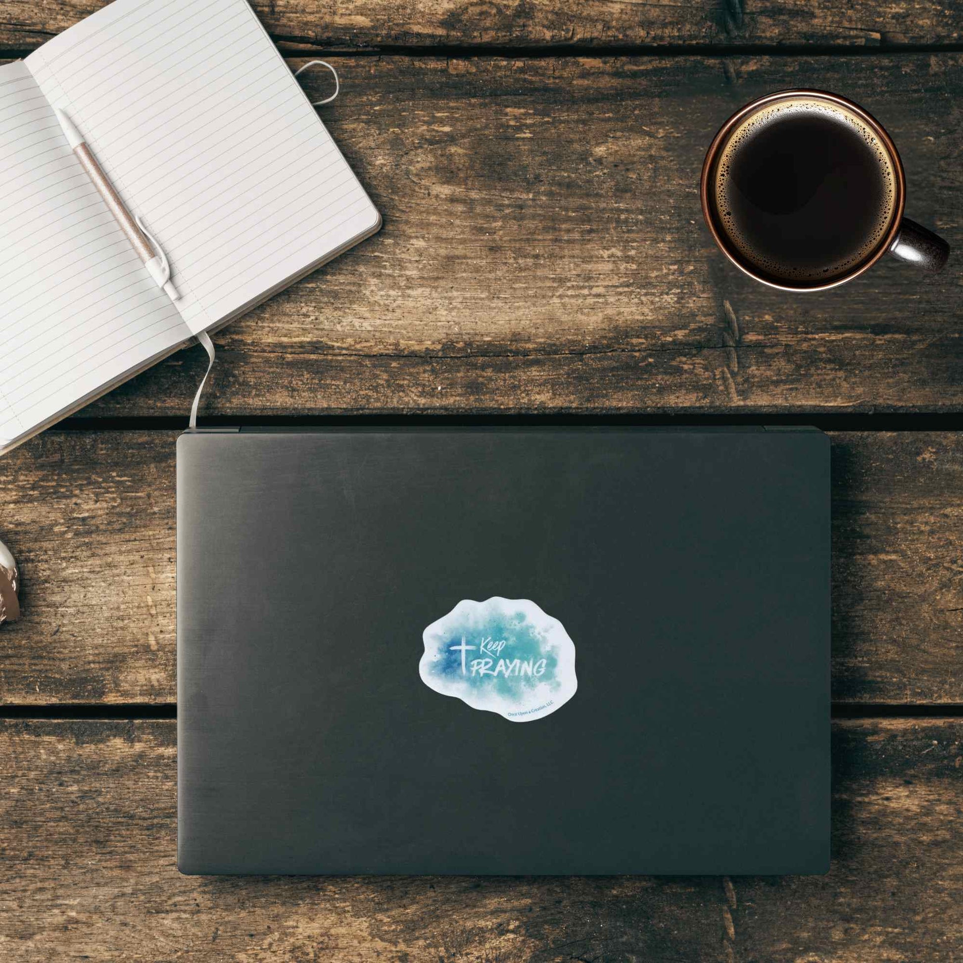 A closed dark gray laptop on a rustic wooden table, featuring a light blue watercolor-style sticker that reads 'Keep praying'. An open notebook with a pen and a cup of coffee are arranged around it in a styled flat-lay composition.