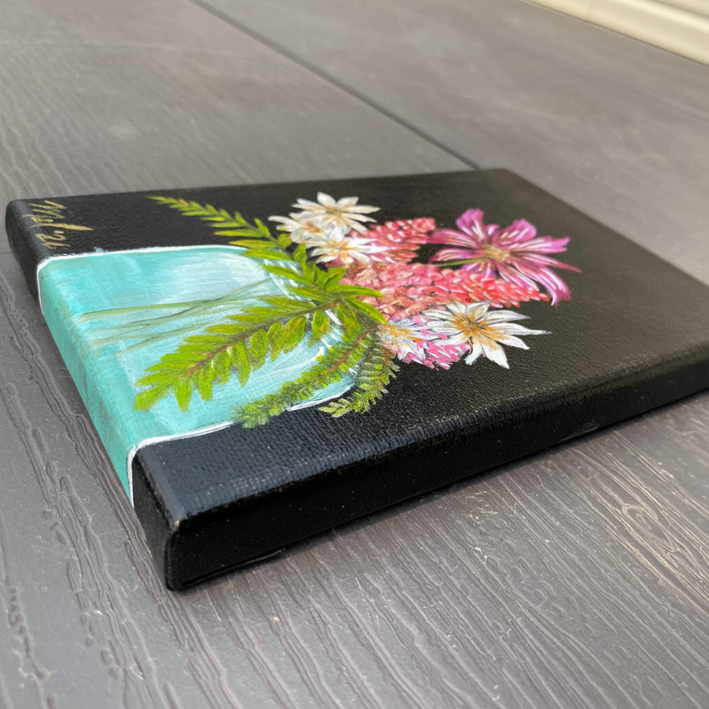 A rectangular canvas painting featuring a painted floral design with pink, white, and green flowers and leaves in a blue mason jar, photographed on a gray wood-textured picnic table.