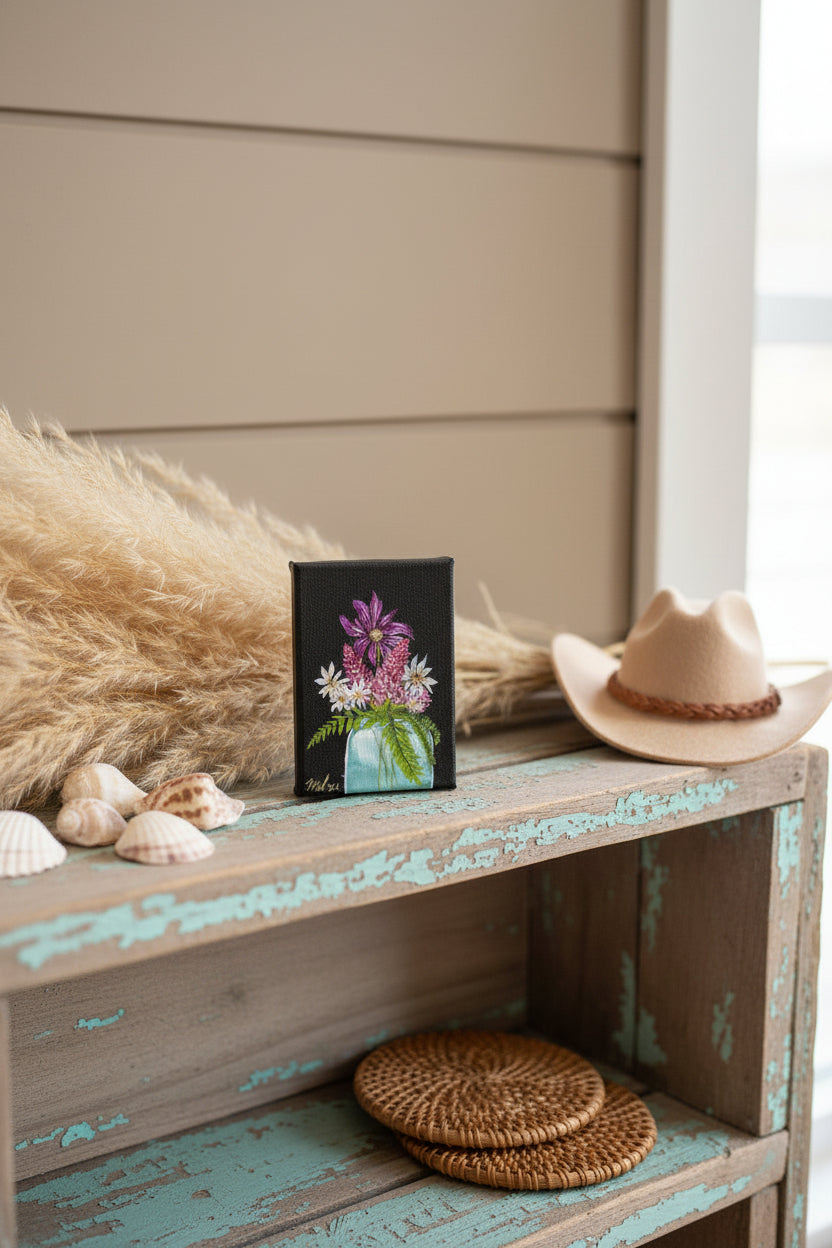 A 5x7" canvas painting featuring a vibrant floral arrangement with purple, pink, and white flowers in a light blue vase against a black background, presented on a coastal cowgirl themed shelf.