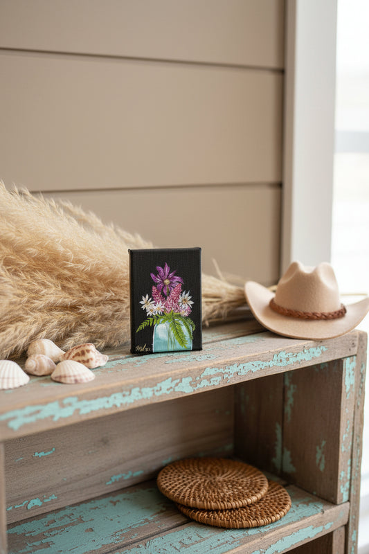 A 5x7" canvas painting featuring a vibrant floral arrangement with purple, pink, and white flowers in a light blue vase against a black background, presented on a coastal cowgirl themed shelf.