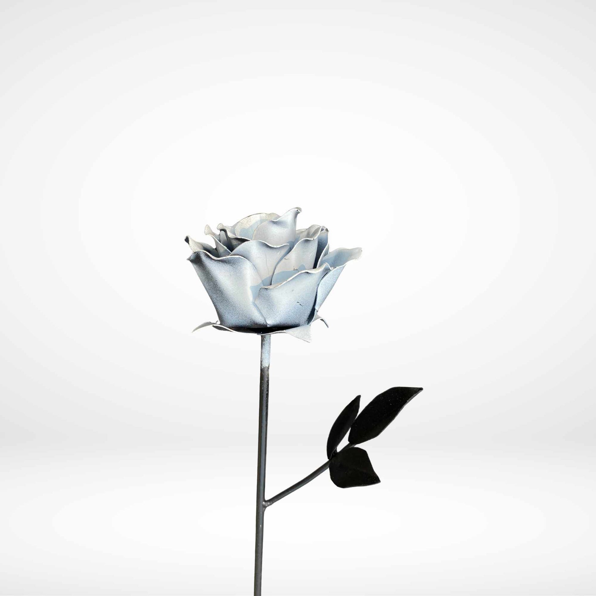 A single metal rose with white petals and black leaves on a long metal stem, photographed against a clean light gray studio background.