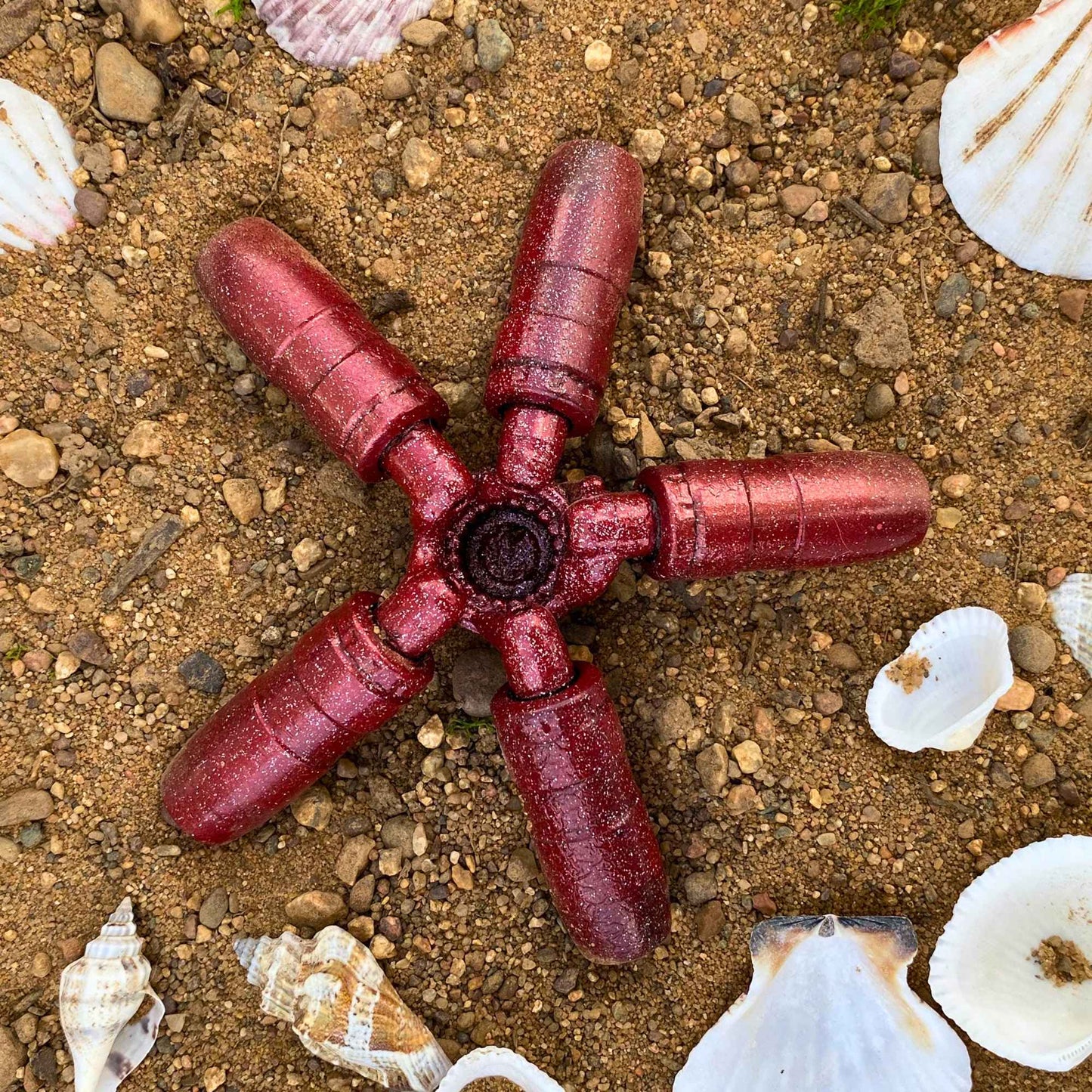 A red sea-star scrap metal sculpture wtih sparkles laying in the sand among seashells. 