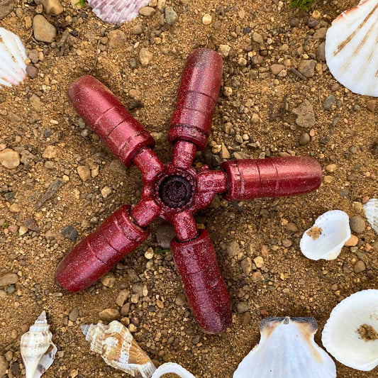 A red sea-star scrap metal sculpture wtih sparkles laying in the sand among seashells. 