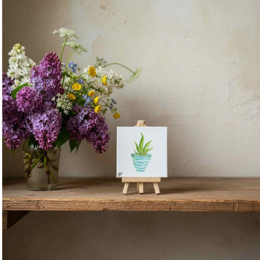 A small square canvas painting of a green aloe plant in a blue pot, displayed on a wooden easel on a rustic wooden shelf. To the left is a glass vase filled with purple, white, and yellow wildflowers against a textured beige wall.