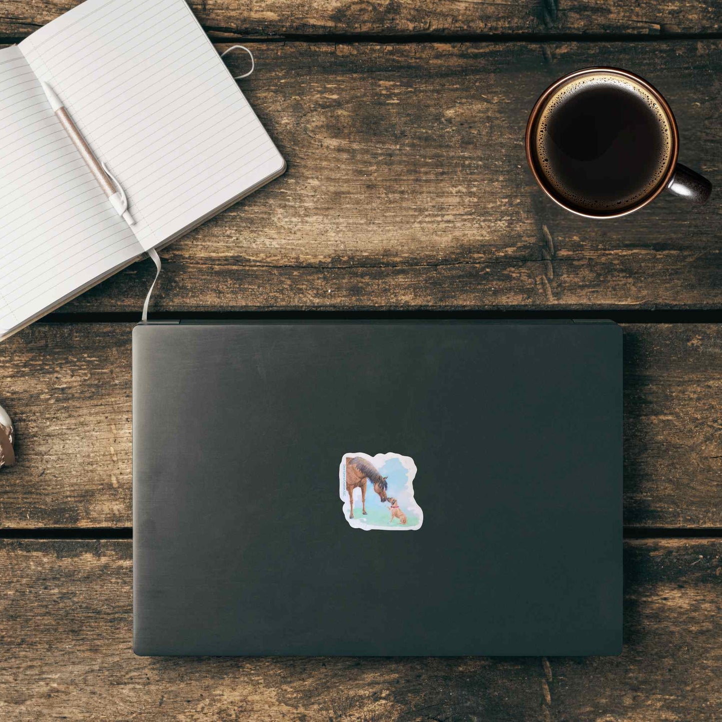 A closed dark gray laptop sits on a rustic wooden table, featuring a small square sticker of a horse being licked by a rescue dog, placed on the lid. An open notebook with a pen and a cup of coffee are arranged around it in a styled flat-lay setting.